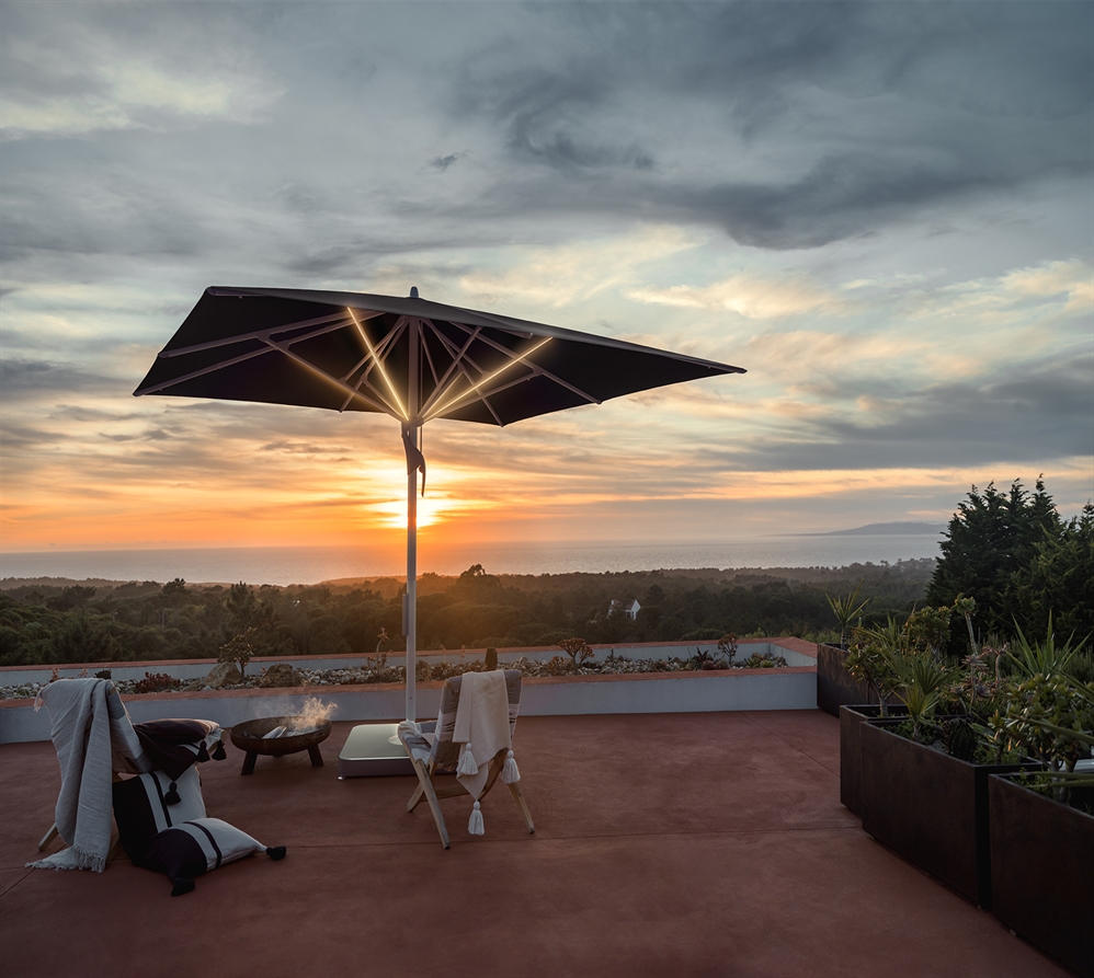 A center pole umbrella with dark fabric and built in LED lighting strips on a patio during sunset