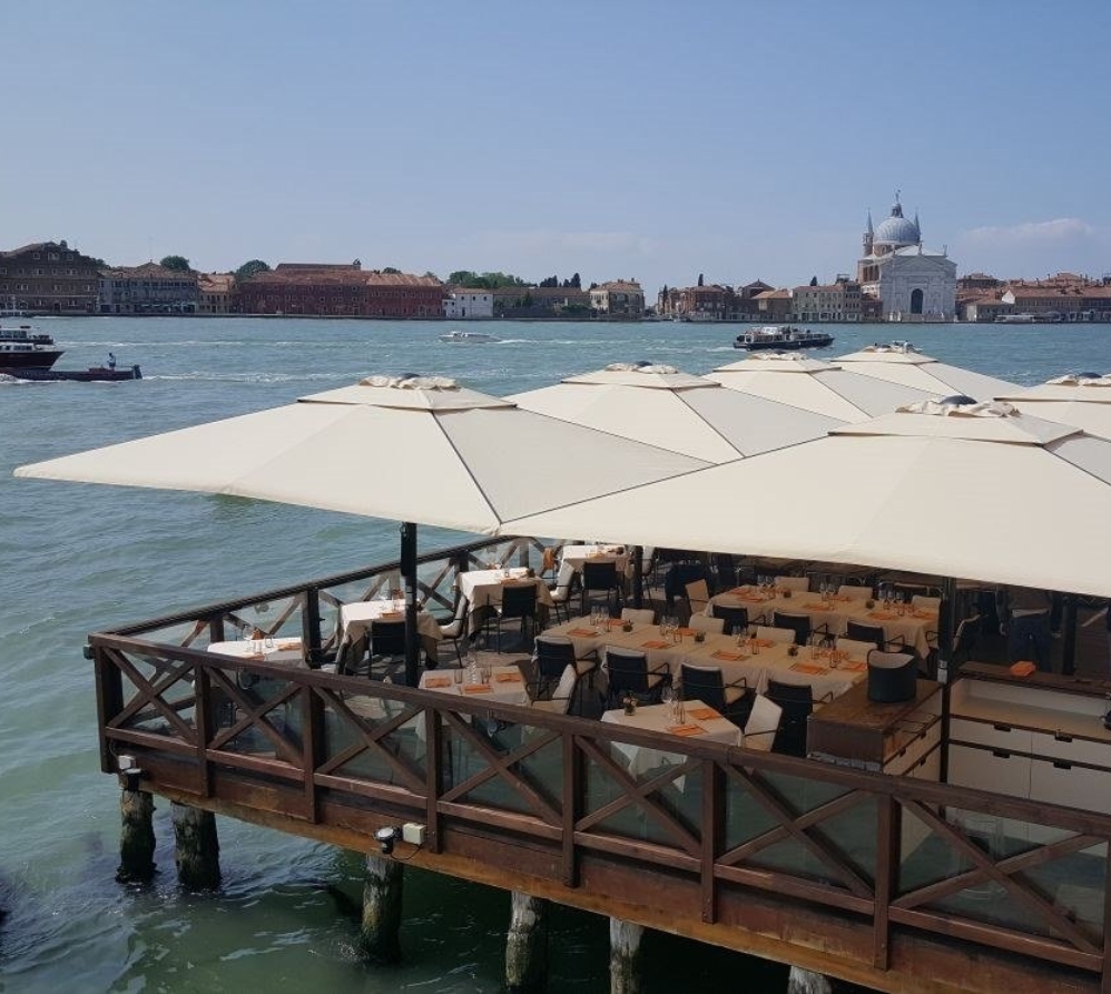 a series of 8 big beige patio umbrella on a patio deck over the water with tables and chairs under
