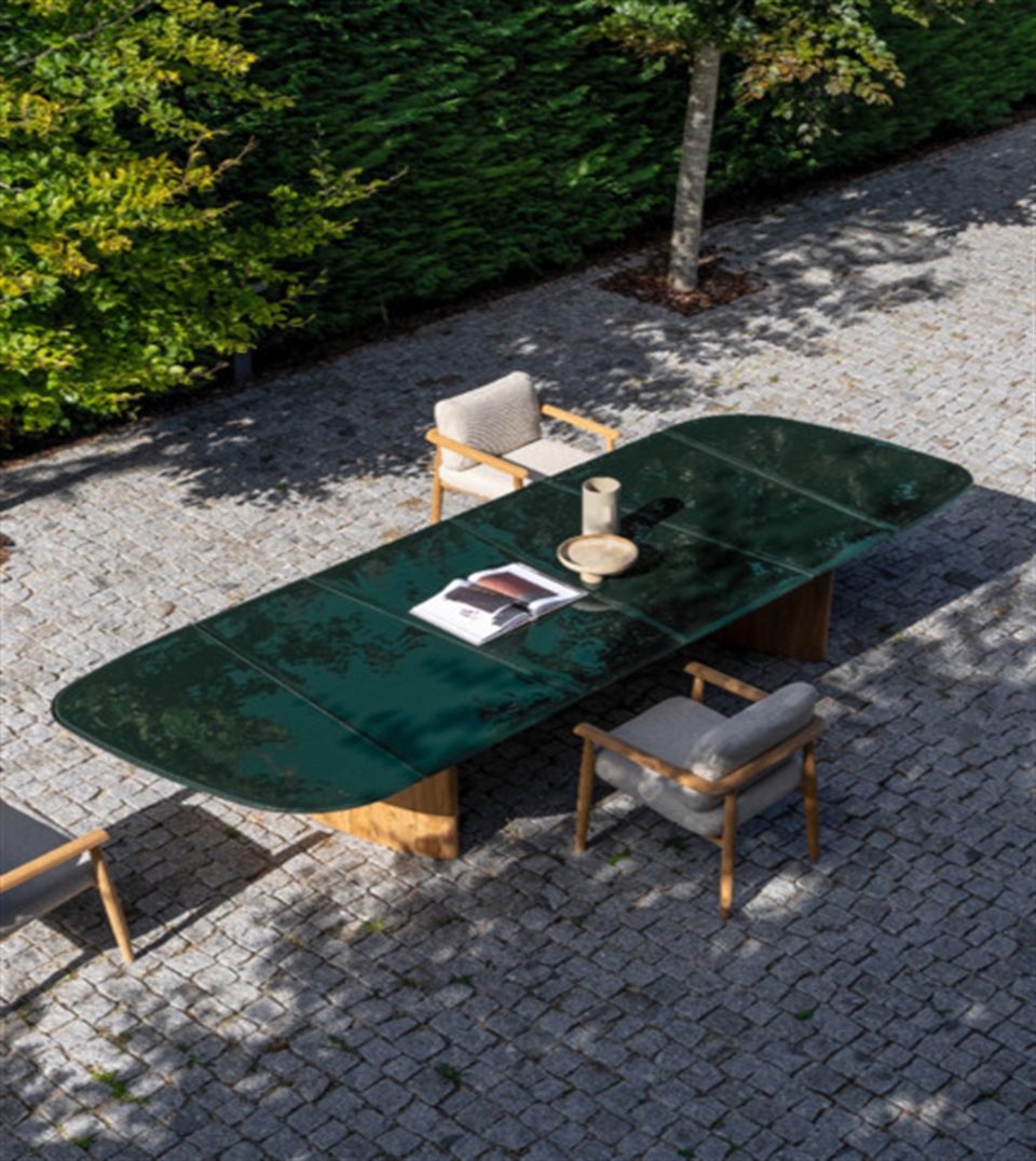 Modern outdoor dining setup with green glass-top table and cushioned wooden chairs on a stone patio.