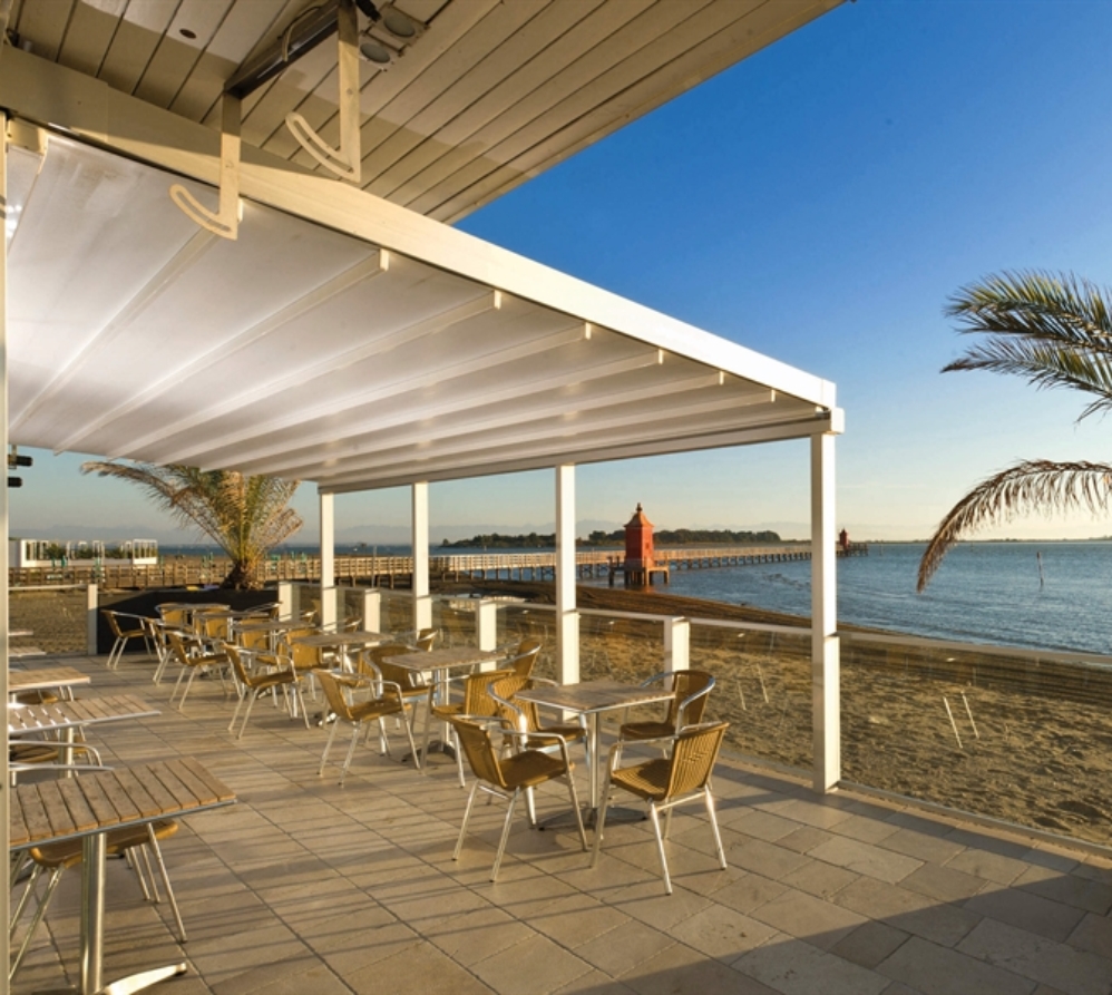 A restaurant patio with a white retractable pvc pergola wall mounted onto the side of the building